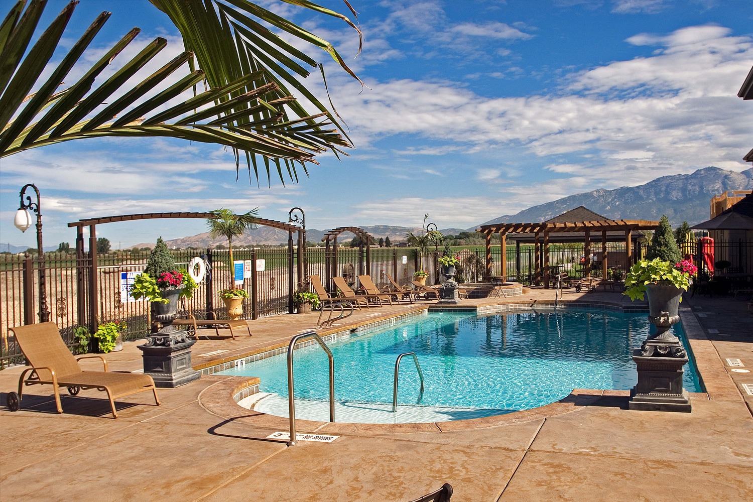 Outdoor swimming pool designed by expert inground pool builders features lounge chairs, potted plants, and pergolas, all set against breathtaking mountain views under a partly cloudy sky.