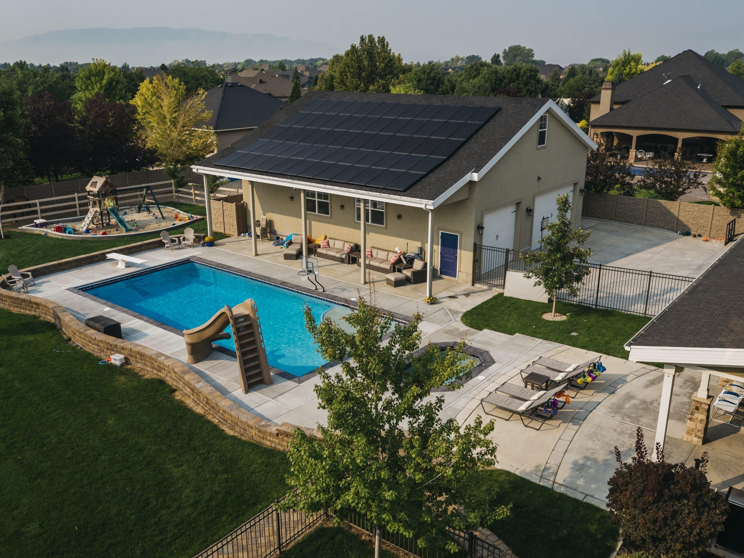 In Sandy Utah, a fenced backyard features a rectangular pool with a slide, lounge chairs, a pool house with solar panels, and a children's playset on the lawn—perfect for relaxing by pools or enjoying family time outdoors.