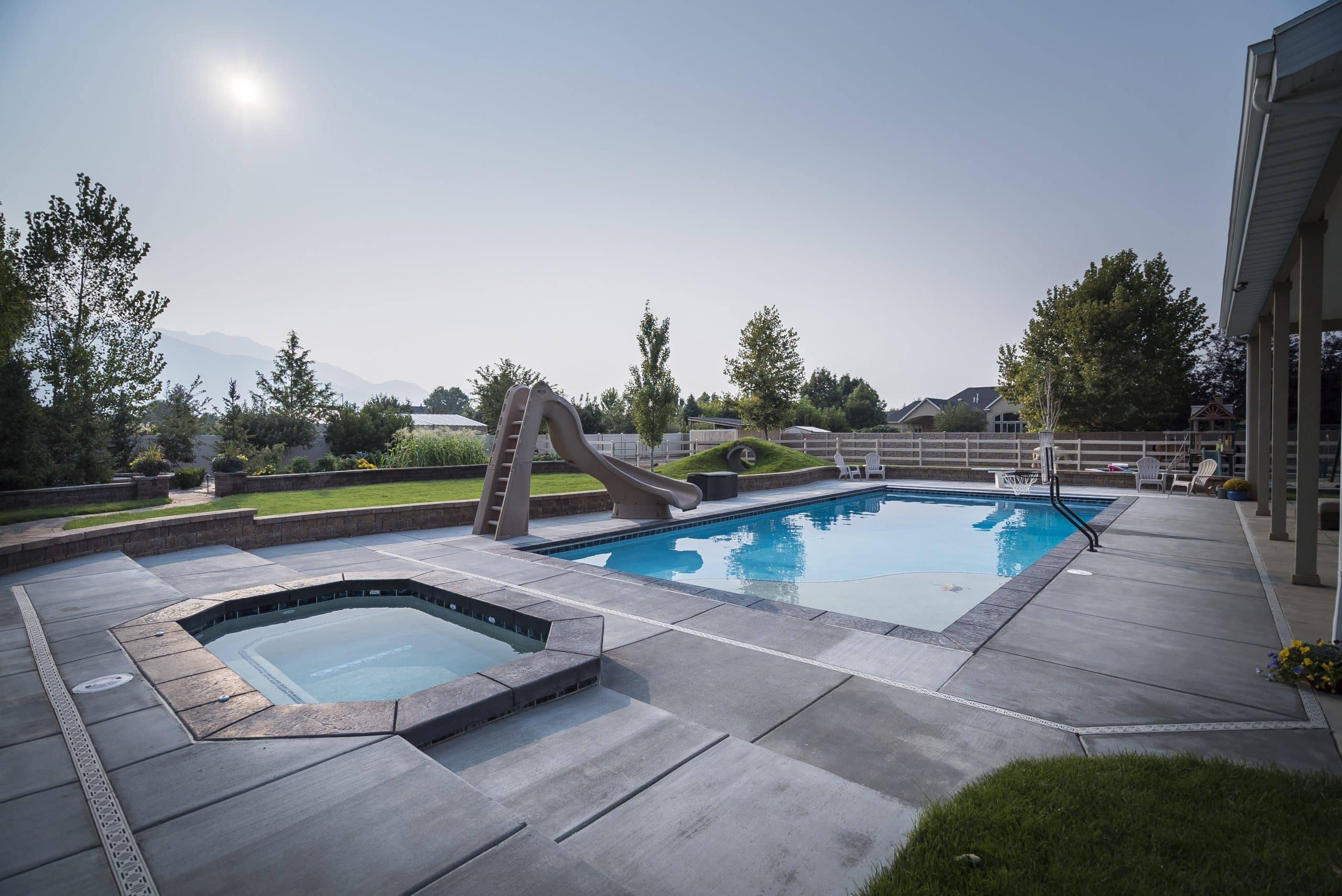 Enjoy a sunny day in your Sandy, Utah backyard featuring an in-ground pool with a slide and adjacent hot tub, all surrounded by a spacious concrete patio. Discover the perfect blend of pools and hot tubs for your outdoor oasis.