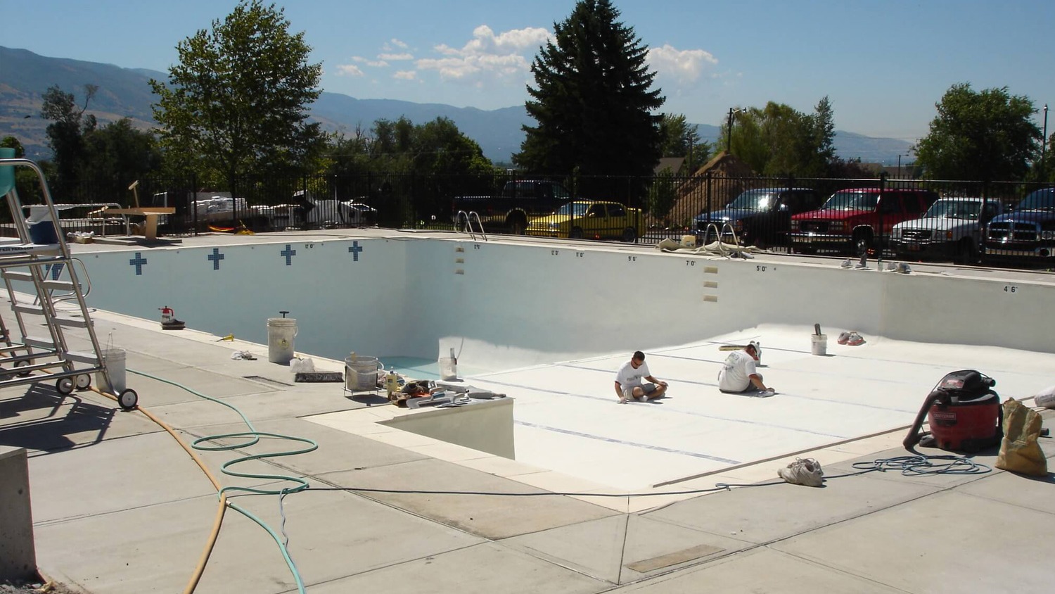 Two workers apply a coating to the interior of an empty inground pool surrounded by construction materials and equipment on a sunny day.