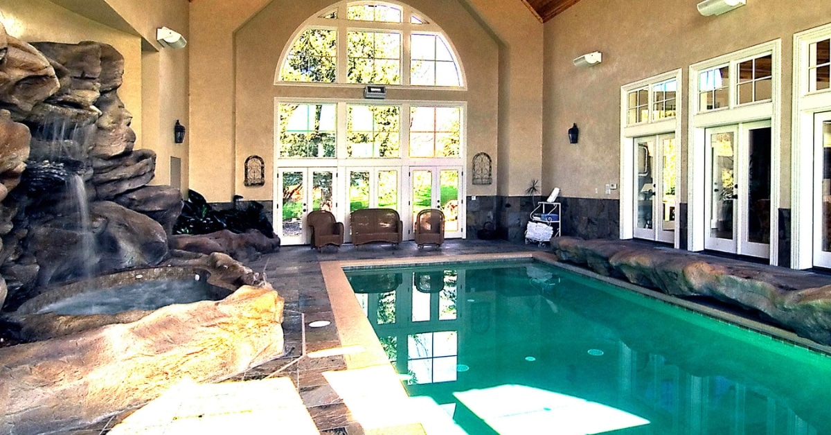 Enjoy a luxurious pool experience with an indoor oasis featuring a natural rock waterfall on the left. Surrounded by large windows and light-colored walls, sunlight floods the area, highlighting chairs perfectly arranged for relaxation by the wall.