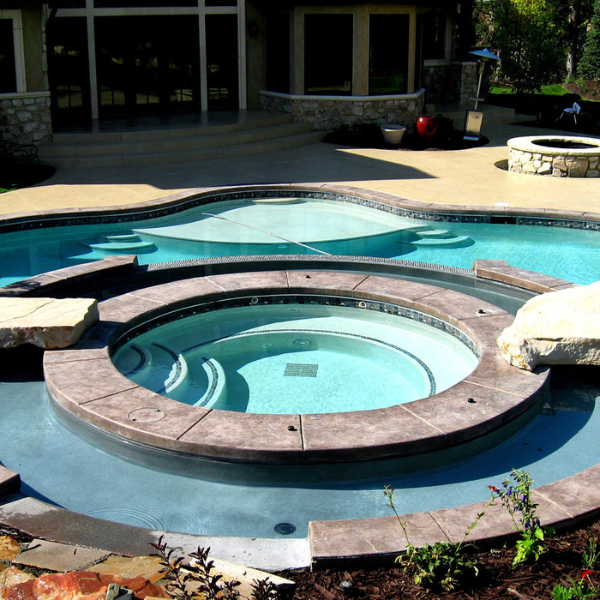 A modern backyard in Sandy UT features an in-ground pool with a circular custom hot tub at the center, surrounded by stone accents and lush landscaping.
