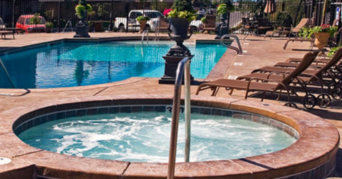 A hot tub in the foreground with bubbling water offers year-round relaxation, adjacent to a larger swimming pool surrounded by lounge chairs and potted plants.