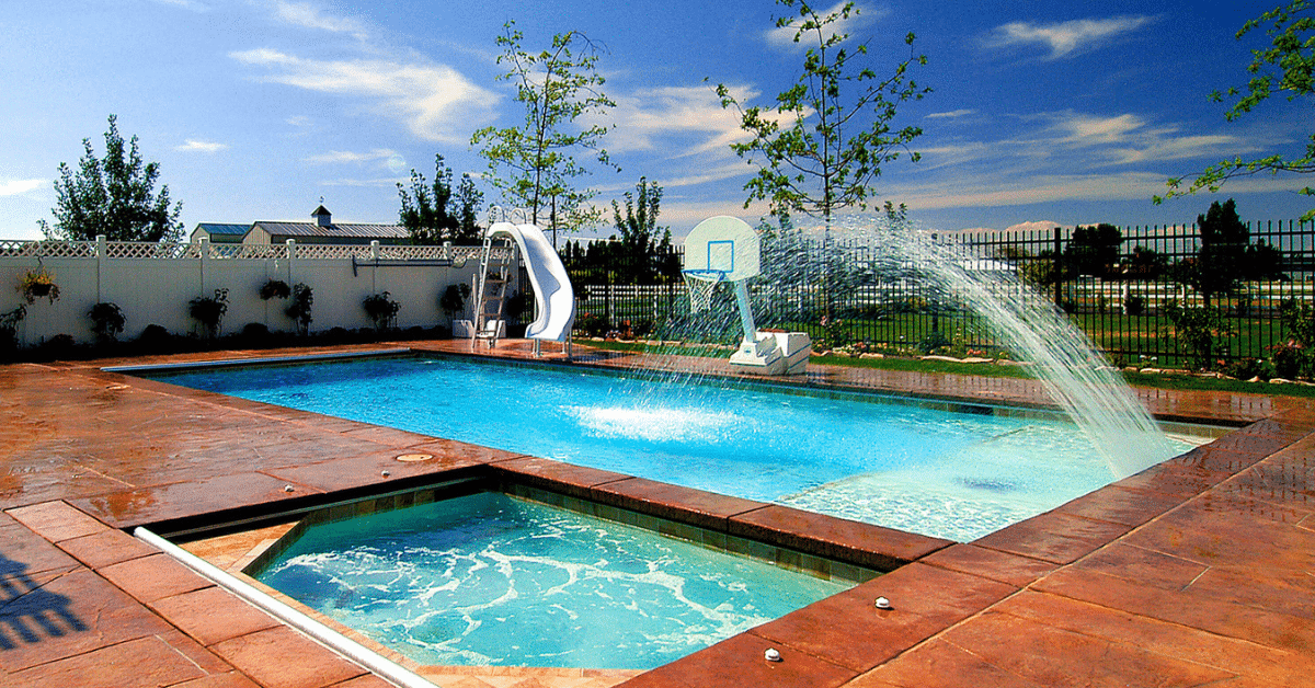 Outdoor pool with a slide and water basketball hoop, incorporating top-notch pool maintenance services. A small adjacent hot tub is in the foreground, surrounded by a concrete patio and fenced by greenery, under a clear blue sky.