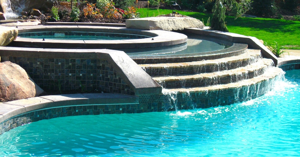 A round stone hot tub overflows into a swimming pool with cascading steps, surrounded by a lush garden, offering a transformative experience.