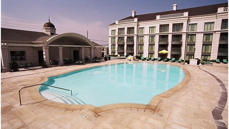 Outdoor hotel swimming pool with curved edges, surrounded by lounge chairs and umbrellas, in front of a multi-story building under a clear sky—expertly cared for with professional pool maintenance so busy homeowners can simply relax and reclaim your weekends.