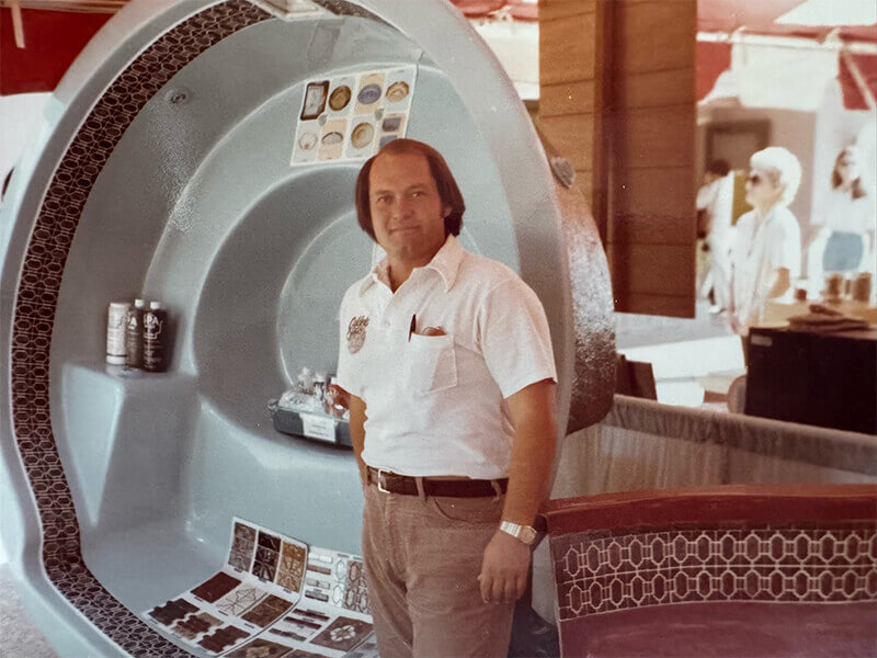 A man in a white shirt stands in front of a large, blue, shell-shaped display booth with various samples and brochures on display.