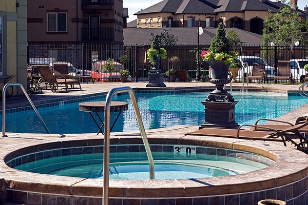 The Aquatech Gallery features a stunning swimming pool area with a hot tub in the foreground, surrounded by lounge chairs and decorative plants. In the background, residential buildings create a serene setting for enjoying luxury Hot Tub Spas.