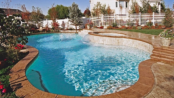 Outdoor swimming pool with clear blue water built by Intermountain Aquatech, featuring an attached circular hot tub, stone decking, landscaped garden, and a white fence in the background—perfect for enjoying the finest in pools and spas.