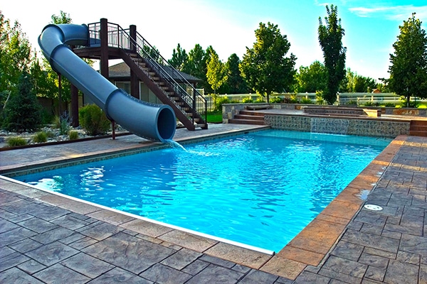 A backyard pool with clear blue water, featuring a waterslide and surrounded by trees and luxurious landscaping, offers a perfect oasis. This stunning example from the Aquatech Gallery creates a serene retreat right at home.