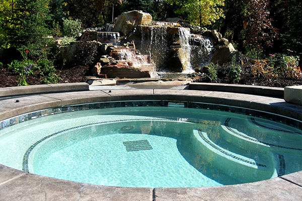 A round, outdoor hot tub spa with clear water is surrounded by stone tiles. In the background, a small waterfall flows over rocks framed by lush greenery, reminiscent of an Aquatech Gallery masterpiece.