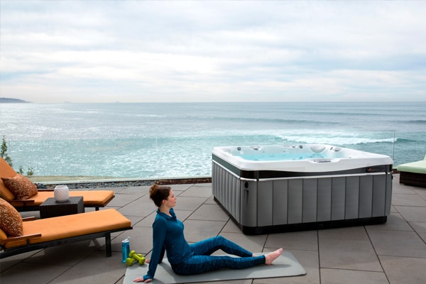 A woman in activewear sits serenely on a yoga mat near an Aquatech Gallery hot tub spa, with an ocean view flanked by lounge chairs and fitness accessories.